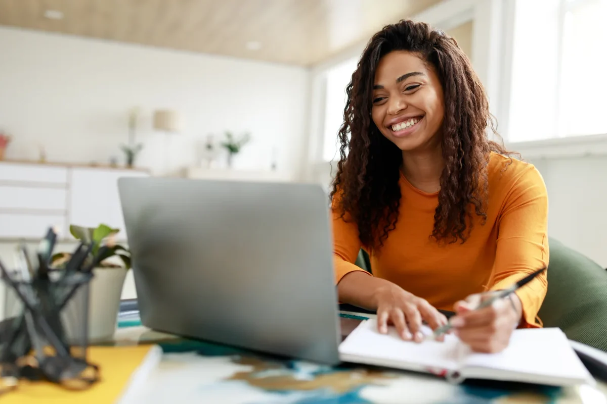 Smiling woman in a yellow shirt using a laptop with an open notebook